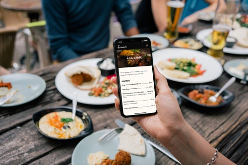 Genrebild på en person som visar sin mobil vid ett matbord. Mobilen visar hemsidan för Frimans i Jönköping som Balowie har varit med och utvecklat.
