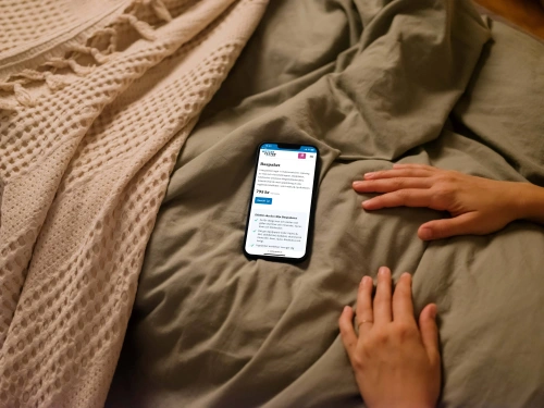 En person som ligger nerbäddad under täcket och har en mobiltelefon lysandes på täcket. Mobilen visar wettertester.se som är en e-handel som Balowie utvecklat.